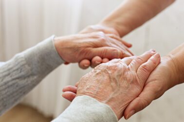 Mature female in elderly care facility gets help from hospital personnel nurse. Senior woman w/ aged wrinkled skin & care giver, hands close up. Grand mother everyday life. Background, copy space.